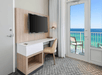 A modern hotel room with a wall-mounted TV, desk, and chair. A glass door opens to a balcony with ocean views.