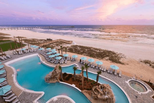 Beachfront resort with a large pool, loungers, and umbrellas overlooking the ocean at sunset. A pier stretches into the water in the background.
