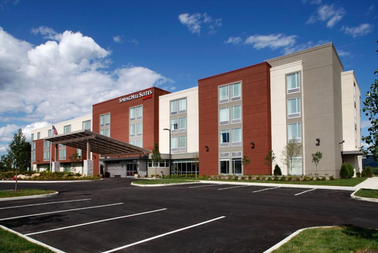A contemporary hotel building with a red and gray exterior, featuring multiple windows and an entrance with a canopy, surrounded by a landscaped parking area.