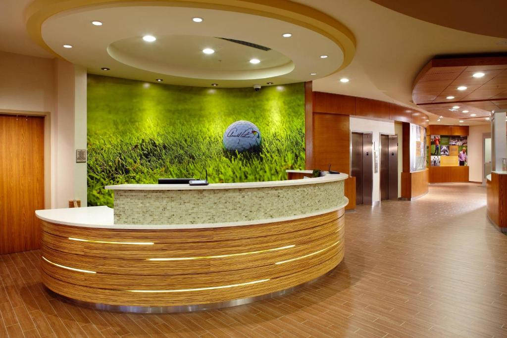 Hotel lobby with a modern wooden reception desk, green golf-themed wall art, and elevators in the background.