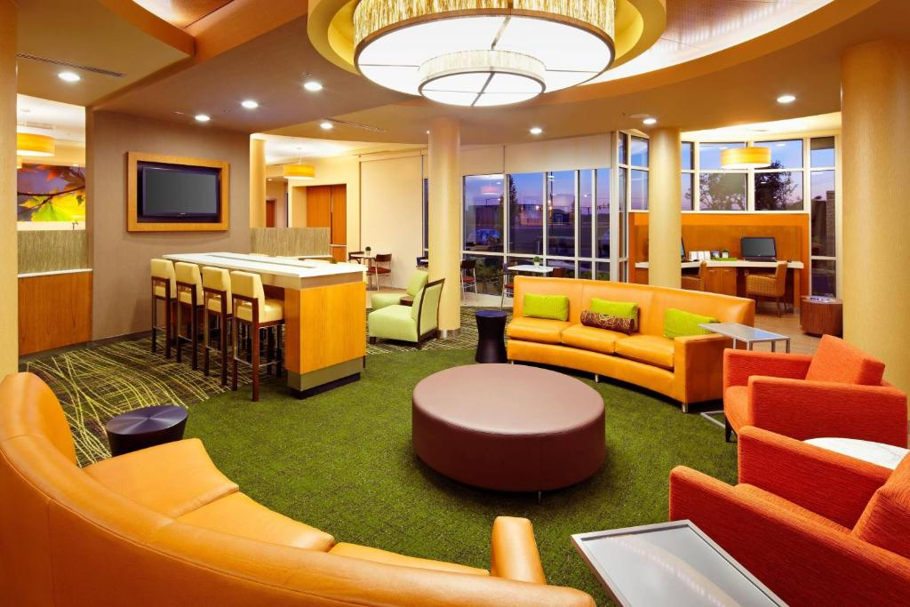 Contemporary lounge area with orange and green seating, a round table, bar stools, and a large overhead light fixture. Large windows provide outside views.