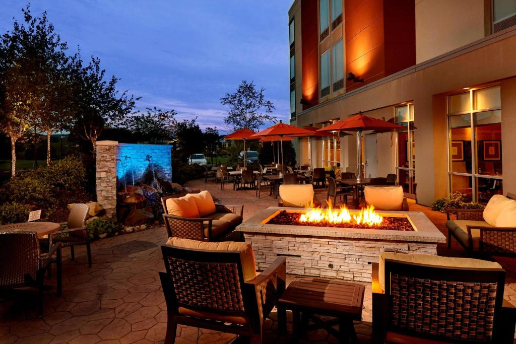 Outdoor patio with a stone fire pit surrounded by cushioned chairs and tables with umbrellas. A small waterfall feature is in the background. The area is lit by building and firelight.