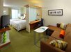 Hotel room with two beds, a sofa, desk, and TV. Decor includes a green carpet, orange accent pillow, and wall art. A frosted glass partition separates the sleeping area.