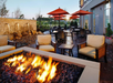 Outdoor patio with a fire pit, cushioned chairs, tables, and red umbrellas. Stone wall and plants are in the background.
