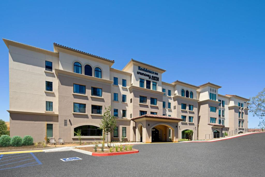 A four-story hotel building with arched doorways, multiple windows, and a flat tile roof under a clear blue sky. The entrance is sheltered by a canopy.