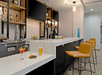 A modern bar setting with a marble counter, featuring drinks, a plate of snacks, and bar stools. Shelves with liquor and two screens are in the background.