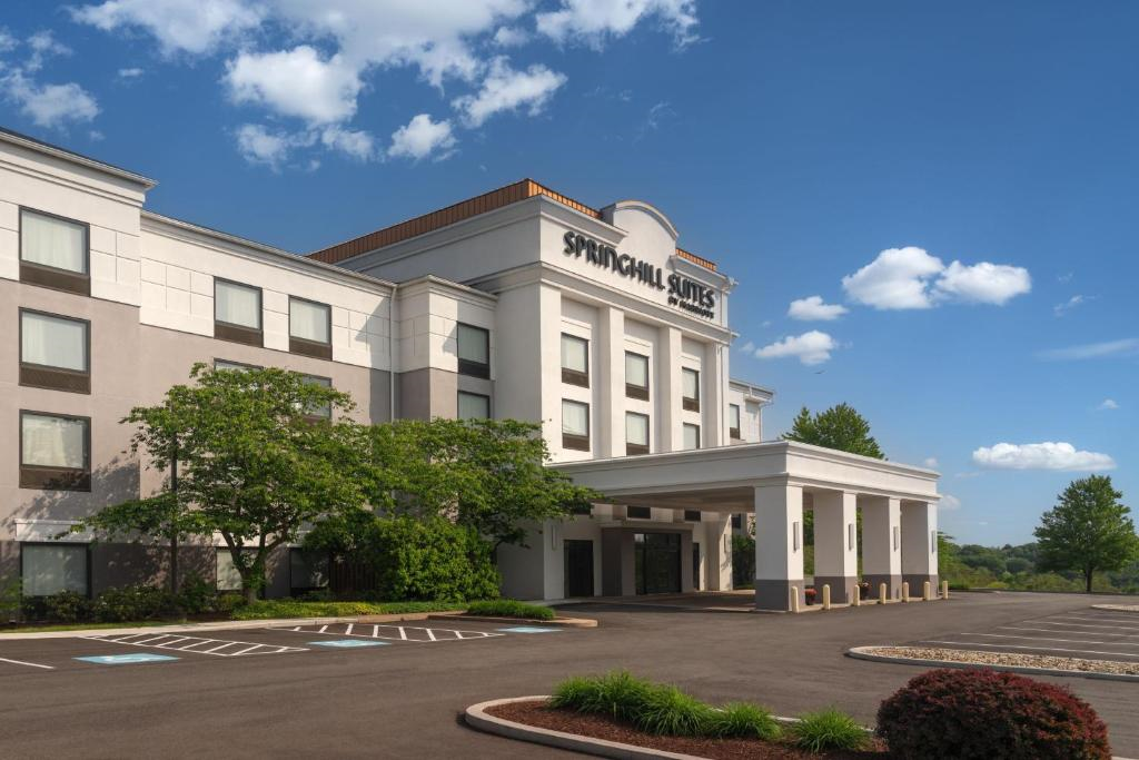 Exterior view of a SpringHill Suites hotel featuring a modern facade, large windows, and a covered entrance. The foreground has a parking area and landscaped greenery under a clear blue sky.