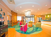 Bright hotel lobby with modern furniture, including red and green chairs, a round decorative light fixture, and a front desk. Wood flooring and a patterned carpet add warmth to the space.