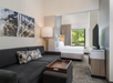 Hotel room with a seating area featuring a dark gray sofa, wooden coffee table, floor lamp, and a bed in the background. A large window provides a view of greenery outside.