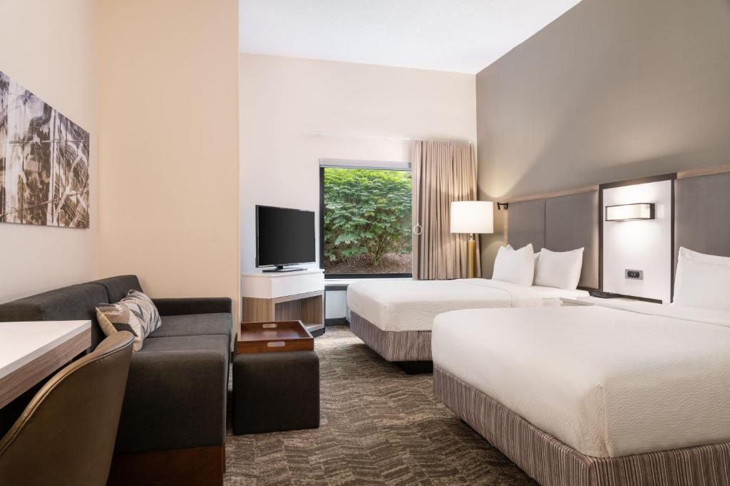 Hotel room with two double beds, a sofa, a TV on a stand, and a desk. Walls are neutral-toned, and a large window shows green foliage outside.