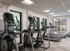 A gym with exercise equipment including treadmills, elliptical machines, and a gray exercise ball. Large windows provide natural light.