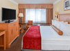 Hotel room with a king-sized bed, red and white bedding, TV on a wooden cabinet, desk with chair, window with curtains, and soft lighting.