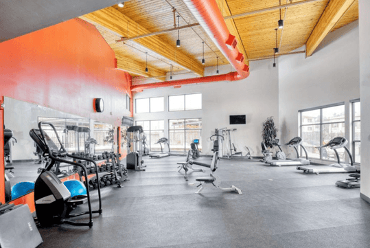 A modern gym with exercise equipment, including treadmills, weights, and stationary bikes. Red ductwork and large windows are visible, providing a bright and spacious atmosphere.
