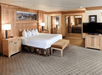 Hotel room with a king-sized bed, two bedside tables with lamps, a bench at the foot of the bed, a flat-screen TV, and a view into the bathroom.