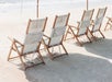 Four wooden beach chairs with white fabric are arranged in a row on a sandy beach under umbrellas.