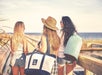 Three women in casual summer outfits walk along a boardwalk toward the beach, carrying bags and wearing sunglasses and hats.
