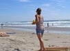 Person in casual attire playing cornhole on a sandy beach, with the ocean and several people in the background.