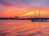 A sailboat is docked near a bridge over calm water at sunset, with vibrant orange and pink clouds filling the sky and a city skyline in the background.