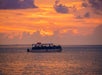 A boat with passengers sails on calm water at sunset, with orange and purple clouds in the sky.
