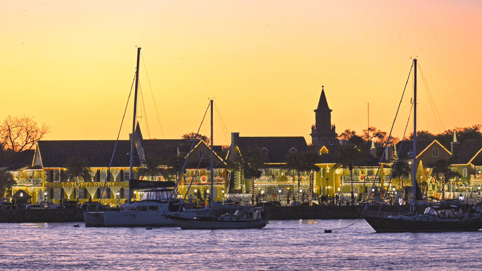 Boats are anchored on the water at sunset with buildings decorated with string lights in the background, and a church steeple visible against the sky.