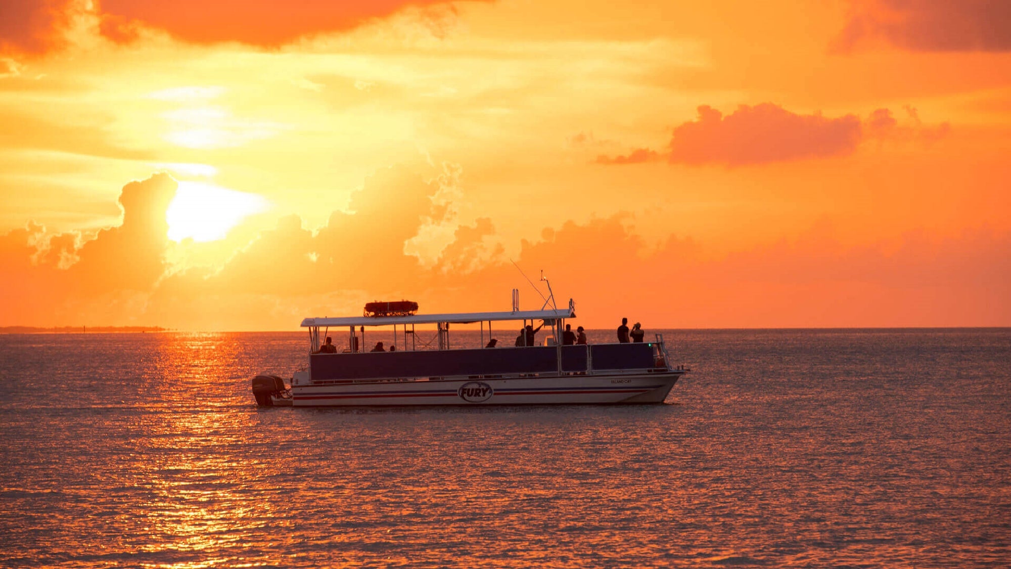 A boat with several people onboard sails on calm water during a vibrant orange sunset.