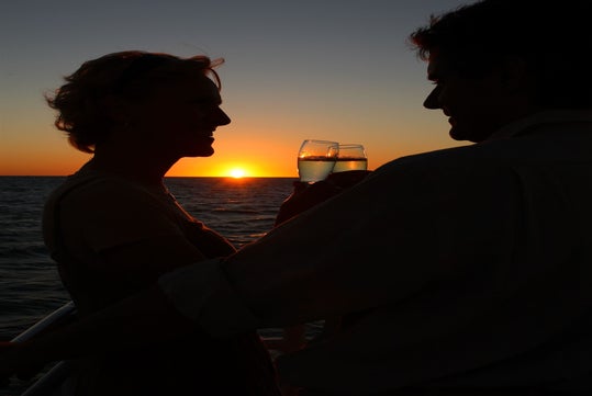 Two people stand close together on a boat, holding glasses and toasting as the sun sets over the ocean in the background.