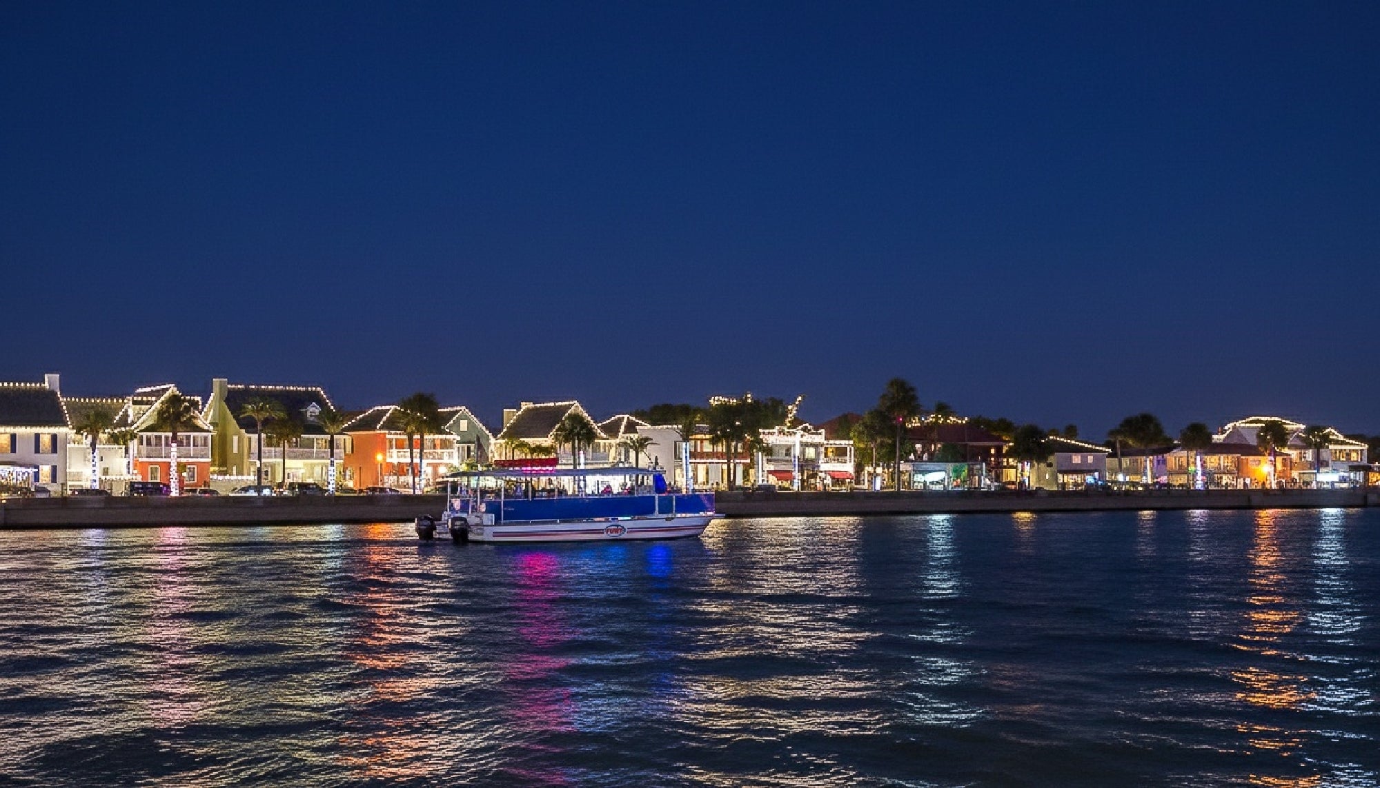 A boat moves across the water at night in front of a row of brightly lit buildings, with colorful reflections visible on the surface.