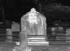 A tall, weathered tombstone stands in the center of a fenced grave plot in a cemetery at night.