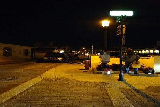 A street musician stands with a guitar near a street corner at night, illuminated by a streetlight. A few pedestrians and a parked vehicle are visible in the background.