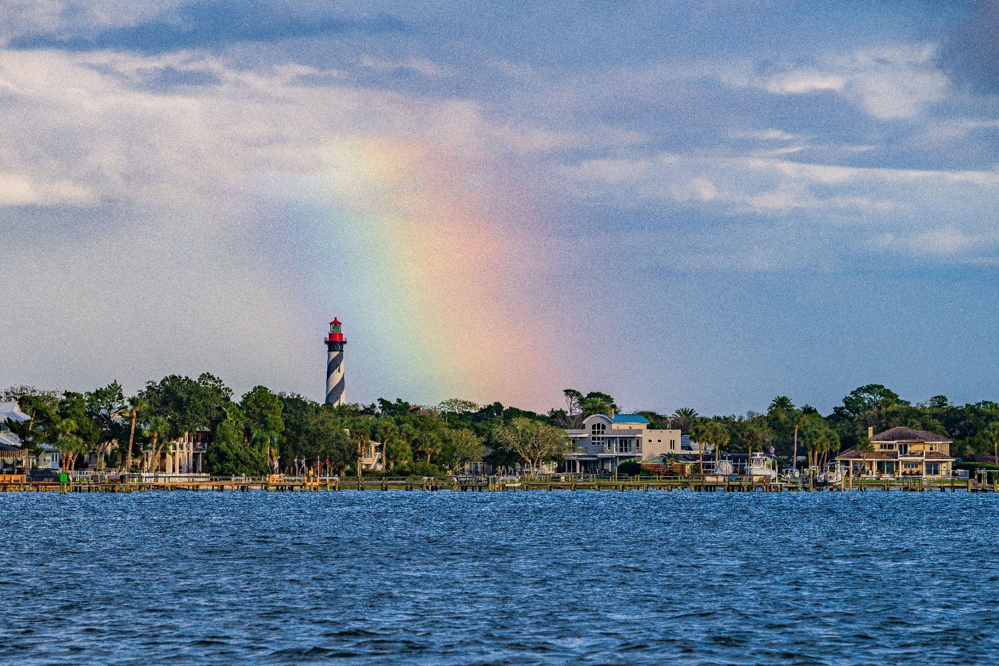 Lighthouse near the shore with a rainbow in the sky, surrounded by trees and houses.