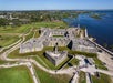 Aerial view of a historic stone fort near a body of water, with surrounding grassy areas and pathways. Visitors can be seen walking around the site.