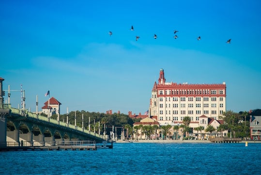 A bridge crosses over blue water toward a historic multi-story building, with several birds flying in the clear sky above.