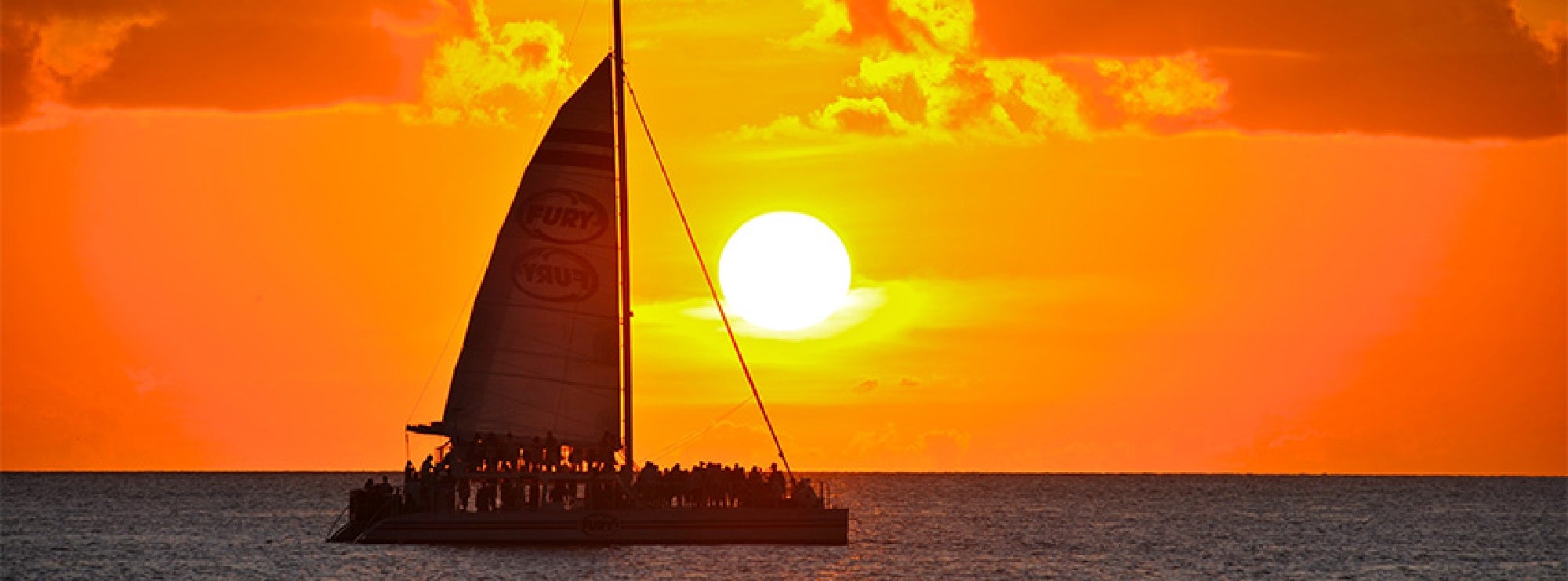 A sailboat with people on board is silhouetted against a vibrant orange sunset over the ocean.