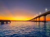 A long bridge with streetlights stretches over calm blue water at sunset, with an orange and yellow sky on the horizon.