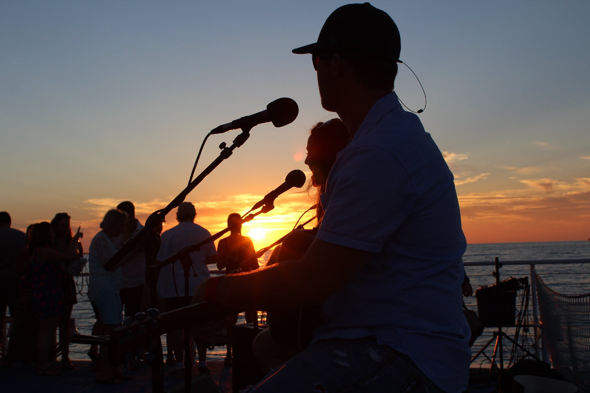 Musicians perform at sunset on a boat deck. Silhouette of a man with a cap playing guitar, with microphones and an audience in the background.