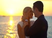 A couple embraces and smiles at each other by the seaside during sunset, with the sun reflecting on the water.