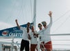 Four people on a boat hold a bottle and wave, standing near a sail with "Signature Fury Charters" text. The sky is clear.