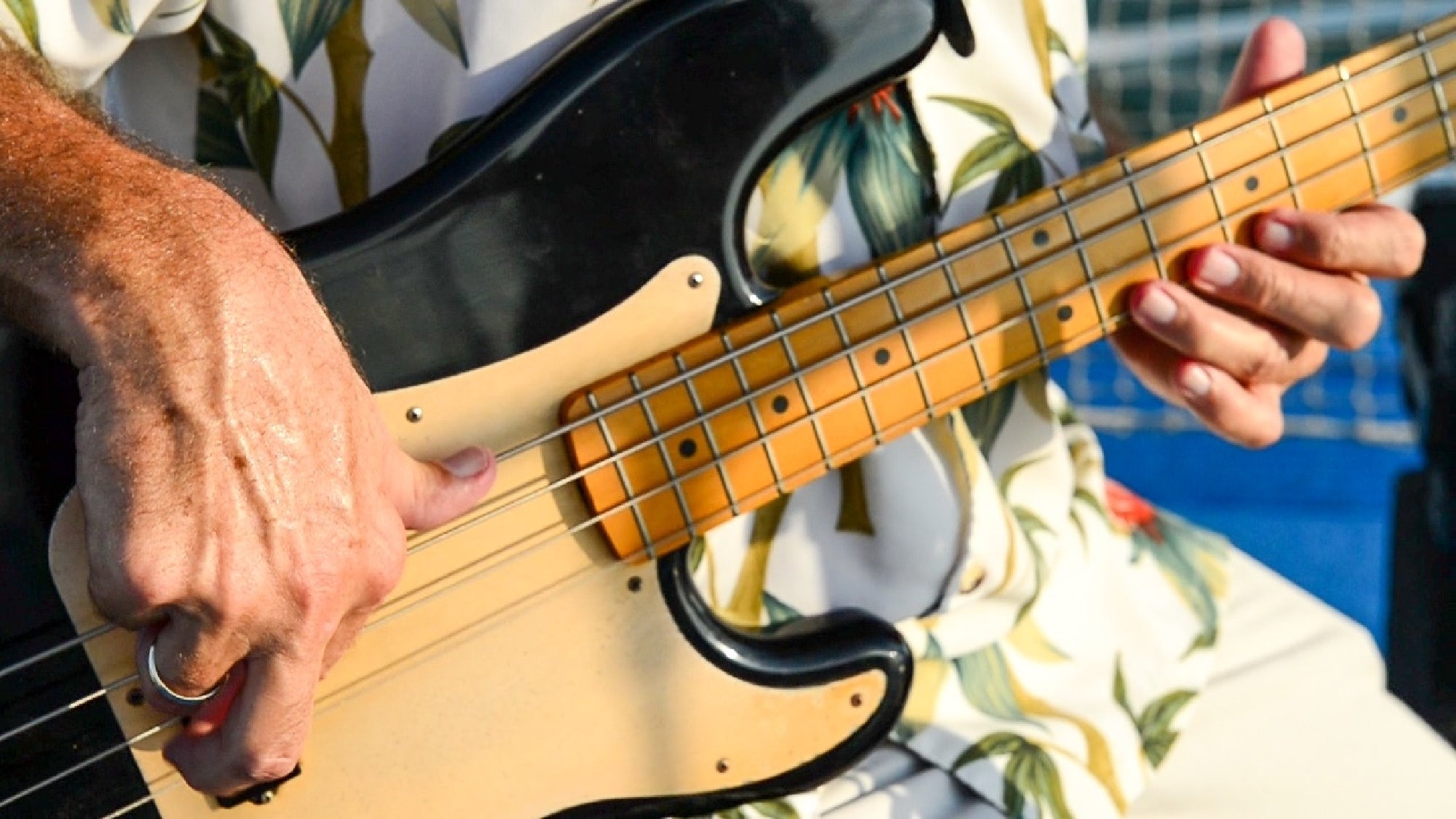 A person in a floral shirt plays an electric bass guitar outdoors.