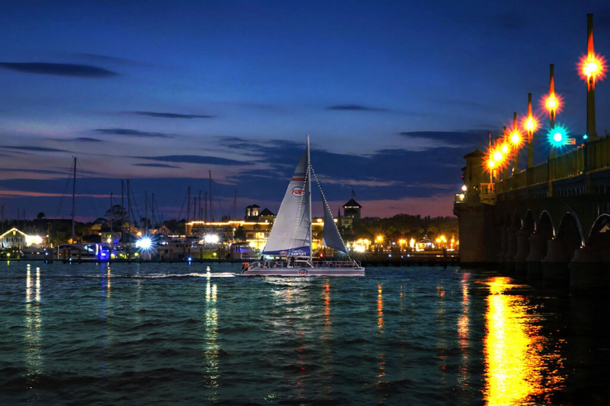 A sailboat glides on the water at twilight near a brightly lit bridge, with a city skyline in the background.