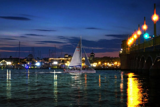 A sailboat glides on the water at twilight near a brightly lit bridge, with a city skyline in the background.