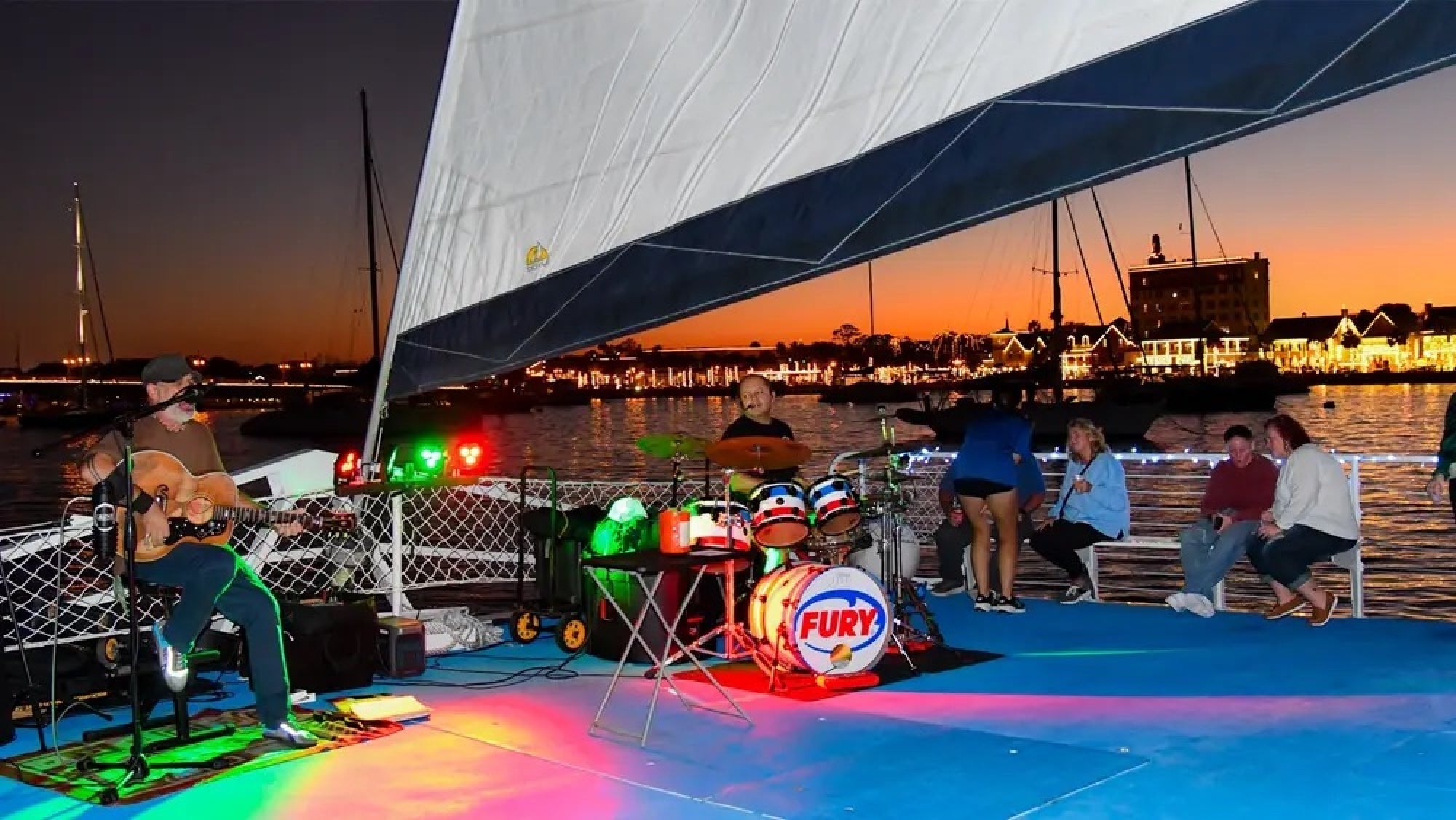 A live band performs on a sailboat at sunset with people sitting and standing on deck, colorful lights, and a harbor visible in the background.