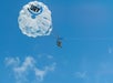 Two people are parasailing high in the sky under a white parachute with "URY" written on it, set against a clear blue sky with a few clouds.