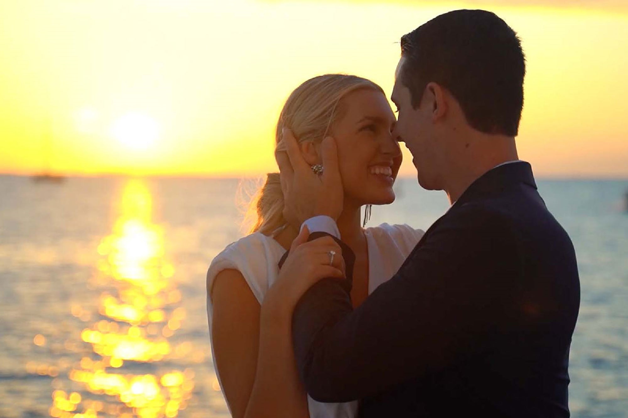 A couple embraces at sunset by the water, with the sun reflecting on the waves in the background.