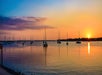 A calm sunset scene over a bay with several sailboats anchored in the water and a vibrant sky displaying shades of orange, purple, and blue.