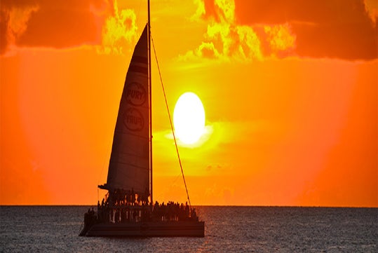 A sailboat with people on board is silhouetted against a vibrant orange sunset over the ocean.