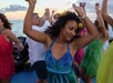 A group of people dancing joyfully on a boat during sunset, with the ocean in the background. The sky is partly cloudy.
