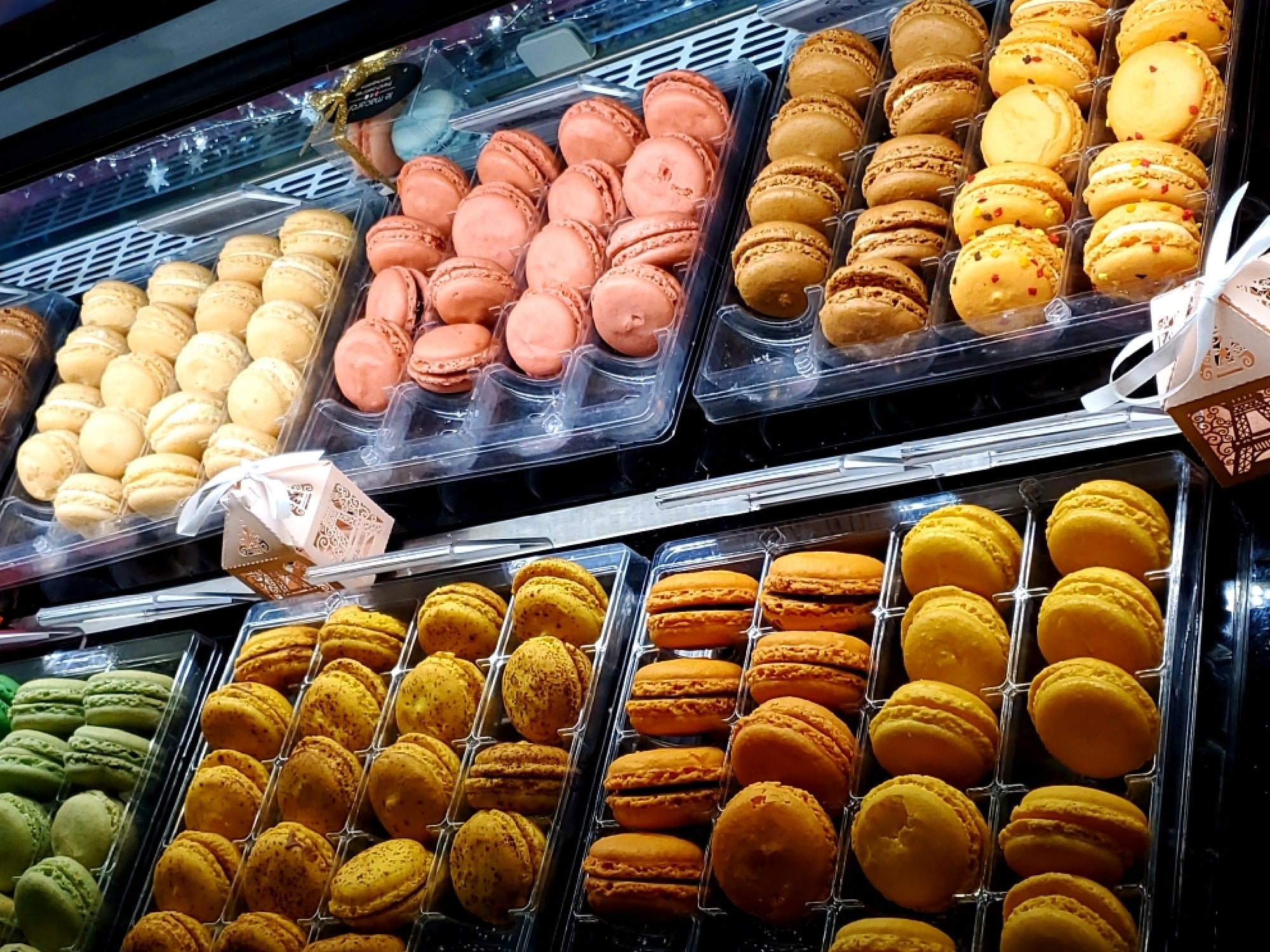 Display case with assorted macarons in various colors, including pink, yellow, and green, arranged in plastic trays.
