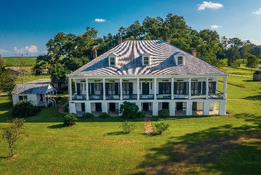 Large historic plantation house with a wide front porch, white columns, and a tin roof, surrounded by green lawn and trees under a blue sky.