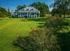 Large white plantation-style house with a wraparound porch sits on expansive green lawn with flowering bushes and trees under a clear blue sky.