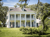 A large white two story plantation style home with black shutters, a second floor balcony along the front, and trees surrounding it.
